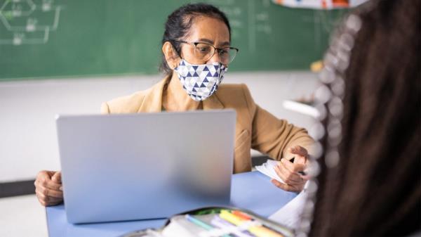A teacher is shown in her classroom wearing a facemask.