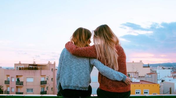 two friends sitting on a rooftop railing, holding each other and looking out at the skyline 